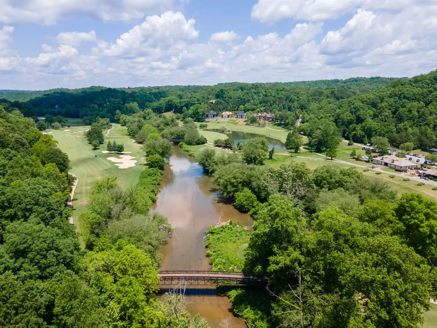 Aerial drone view of the Chestatee River winding through the Achasta golf course in Dahlonega Georgia, with a pedestrian footbridge crossing the river and estate homes nestled in hardwood canopy
