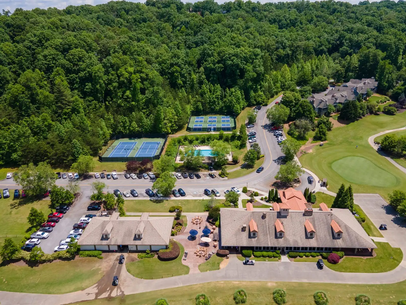 Aerial view of the Achasta clubhouse, tennis and pickleball courts, swimming pool, parking lot, and a manicured golf green