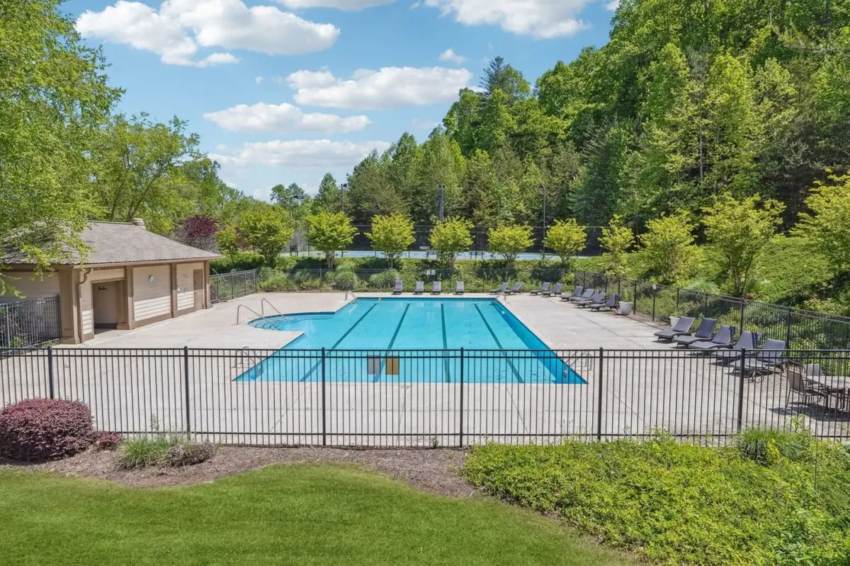 Achasta community swimming pool with lap lanes, dark lounge chairs along the deck, pool house at left, and an evergreen privacy hedge behind