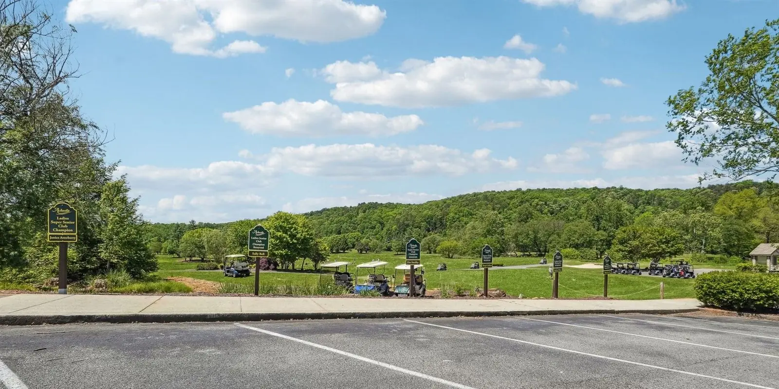 The Achasta practice range and cart staging area seen from the edge of the parking lot, with golf carts parked along the path, signs on posts and a hardwood ridge under a blue sky