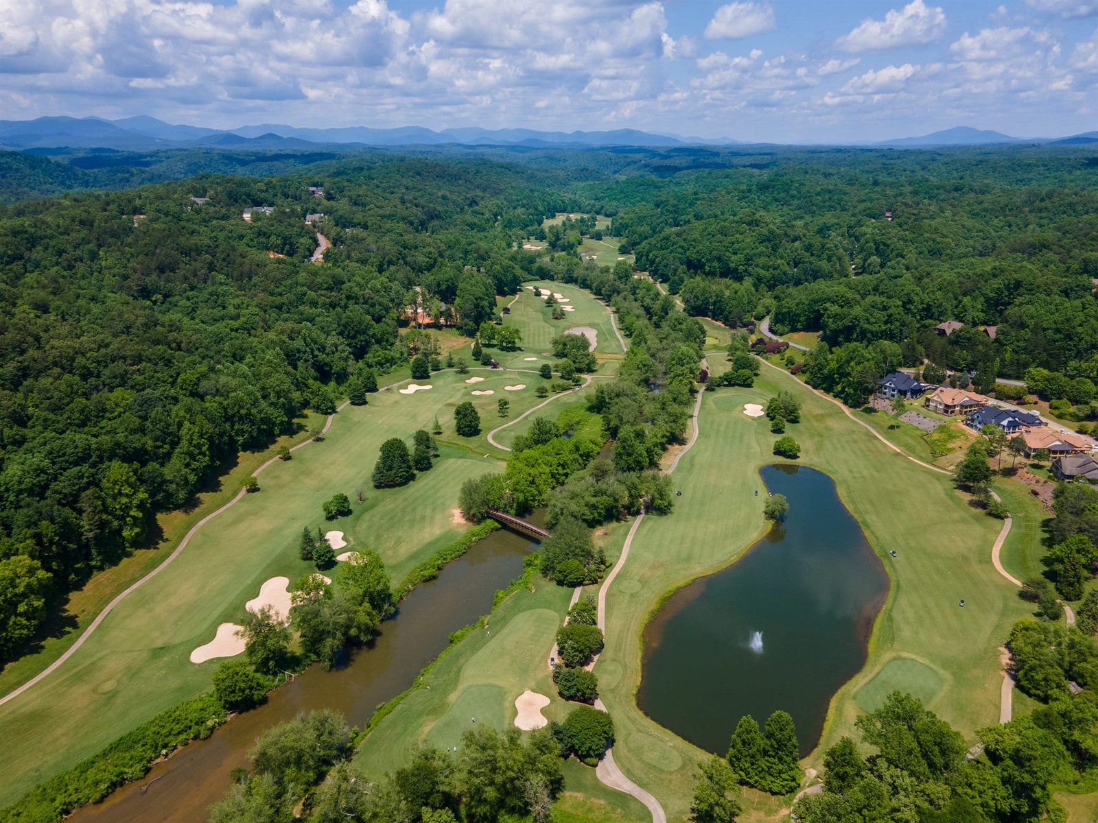 Aerial drone view of the Jack Nicklaus signature golf course at Achasta in Dahlonega Georgia, with a pond and fountain in the foreground and the rounded Blue Ridge Mountains across the horizon