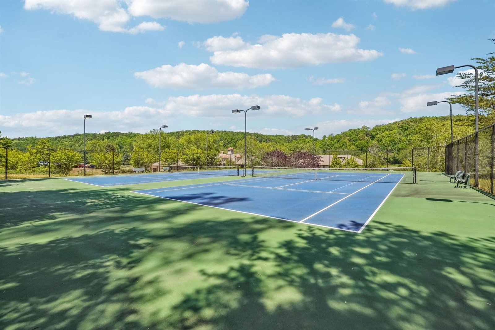 Two hard tennis courts at Achasta with light poles, with the clubhouse and tree-covered hill in the background