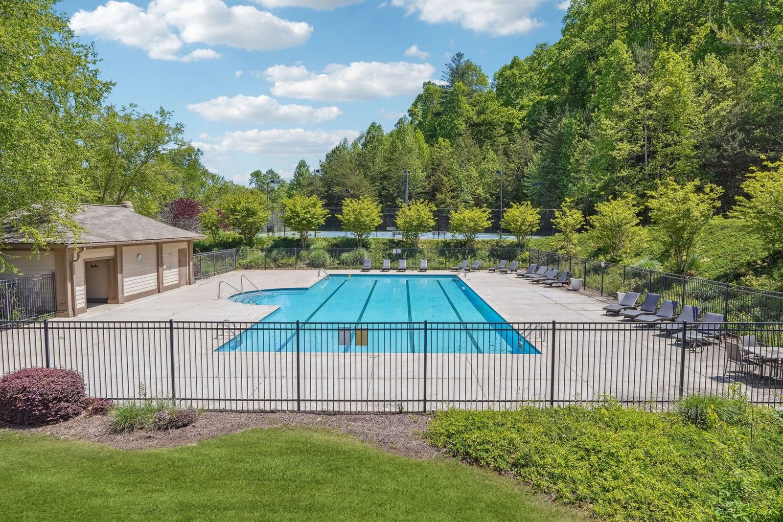 Achasta community swimming pool with lap lanes, dark lounge chairs along the deck, pool house at left, and an evergreen privacy hedge behind