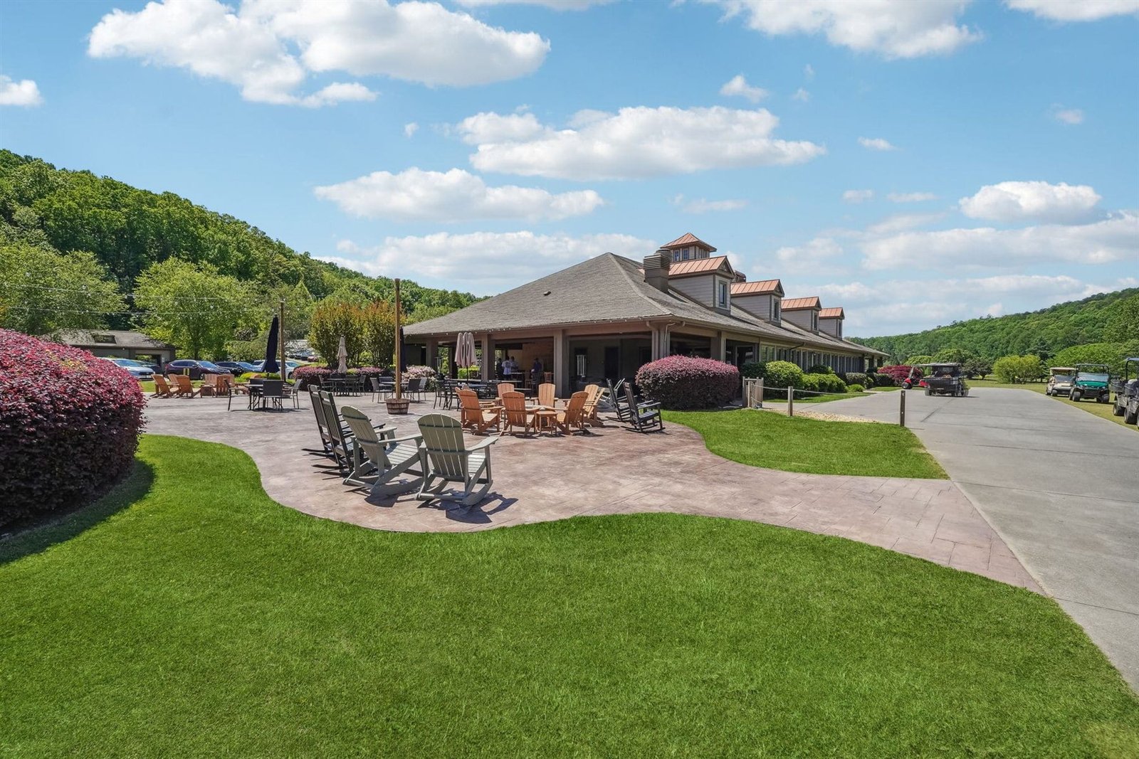 Exterior of the Achasta clubhouse with stamped concrete patio, Adirondack seating, paved cart path, and hardwood-covered hills behind