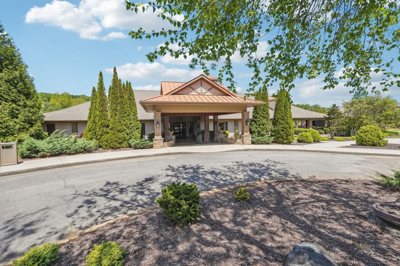 Front entrance of the Achasta clubhouse with copper-toned gable porte-cochère, topiary, and a paved circular drive