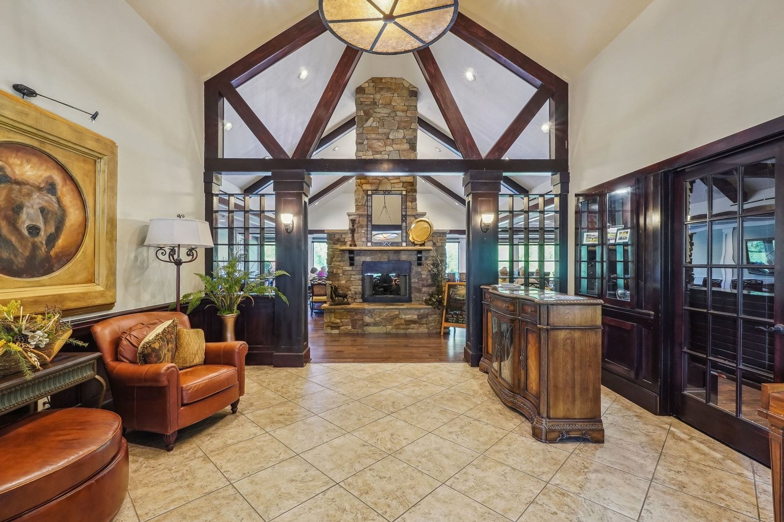 Interior of the Achasta clubhouse great room &mdash; vaulted ceiling with exposed dark timber trusses, floor-to-ceiling stone fireplace with antique clock, leather club chairs, and a bear-portrait painting on the wall