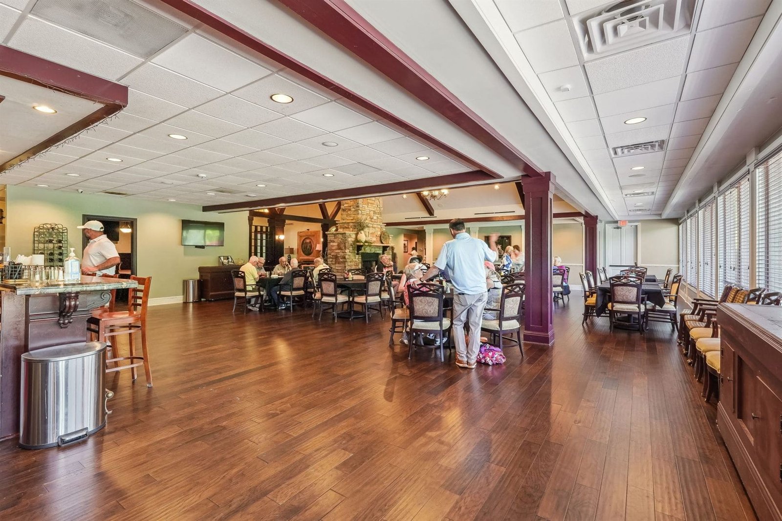Achasta clubhouse dining hall with hardwood plank floors, members seated at dining tables, and a fireplace visible in the room beyond