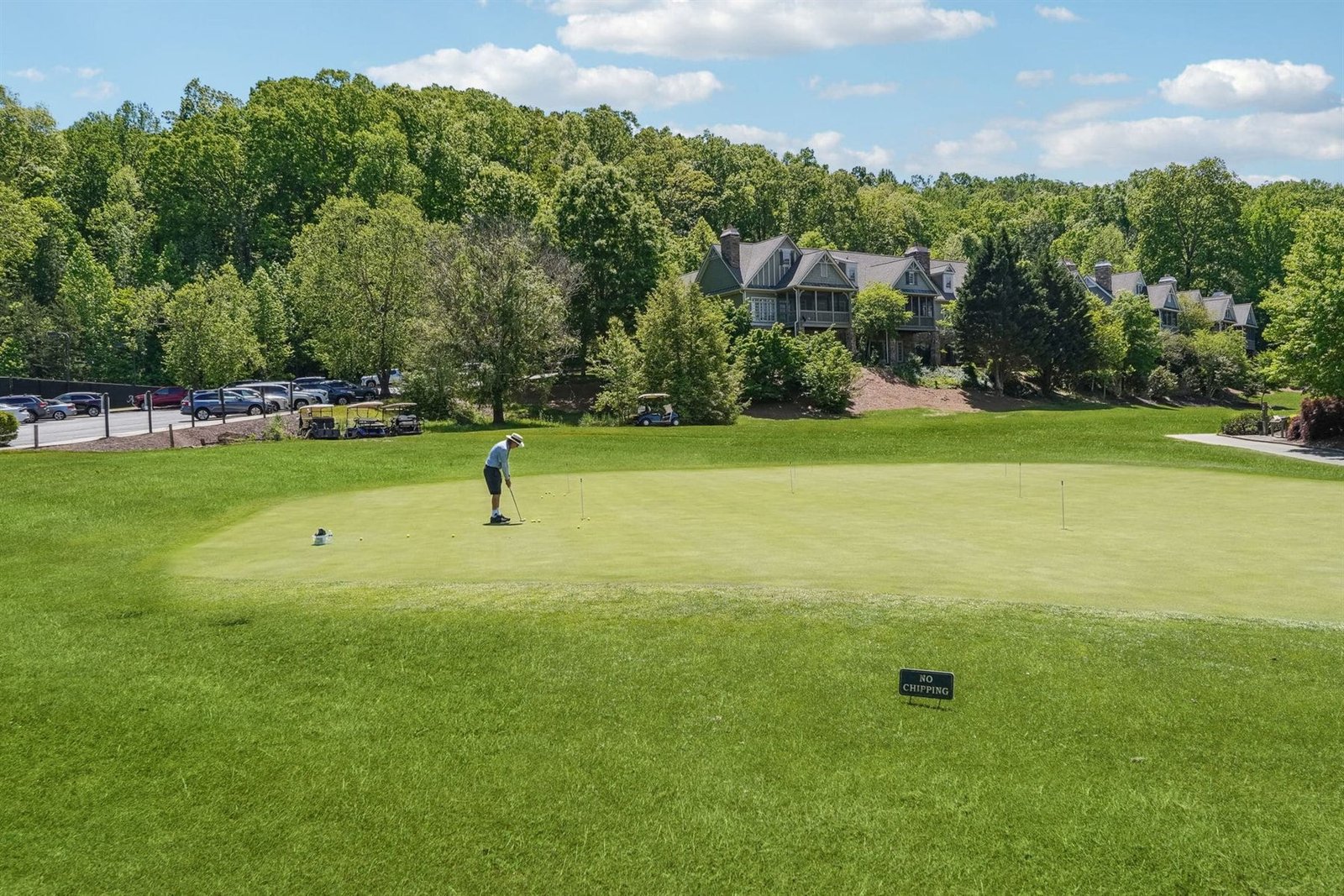 A single golfer practices on the Achasta putting green with a large gabled stone and shake estate home in the background