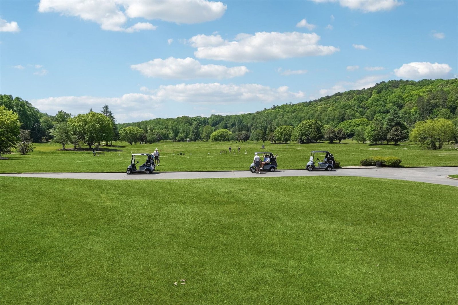 Four golf carts traveling along a paved cart path that crosses a fairway at Achasta with a hardwood ridge across the horizon