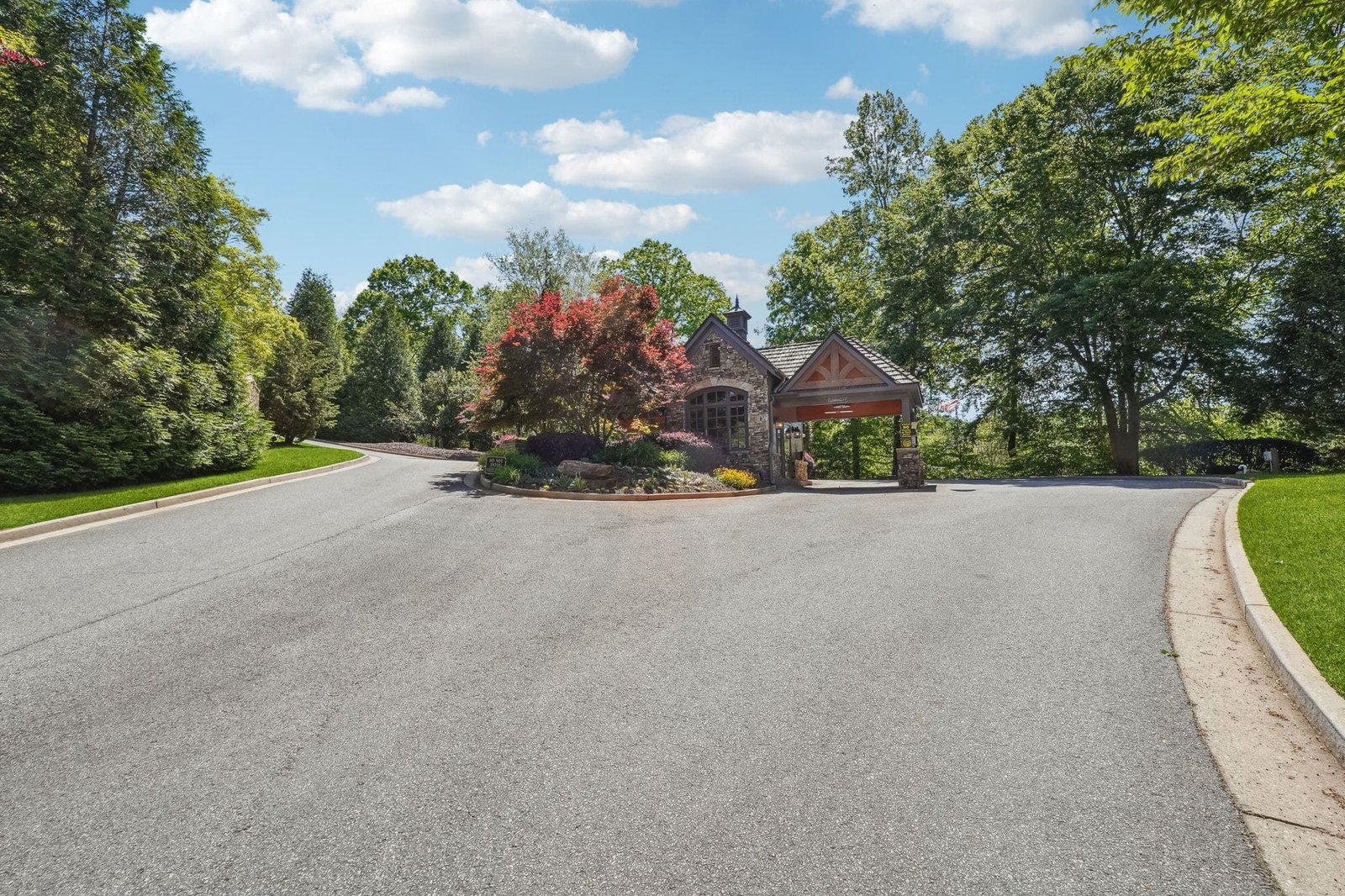 Achasta primary gate &mdash; stone gatehouse with timber porte-cochère for the gate keeper, ornamental landscaping with red-leaf shrubs in the median, and a wide paved entry sweep