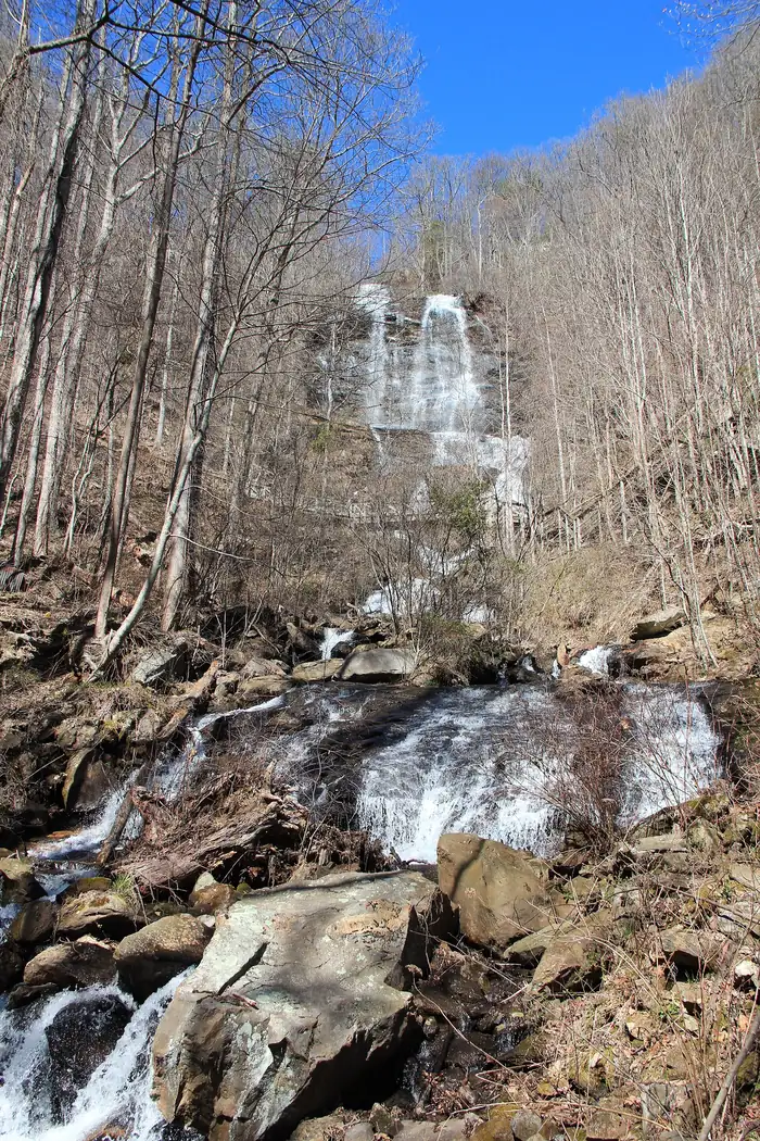 Amicalola Falls in Georgia seen from below in early spring: the upper cascade high on the slope above a rocky lower cascade, framed by bare hardwood trees under a blue sky.